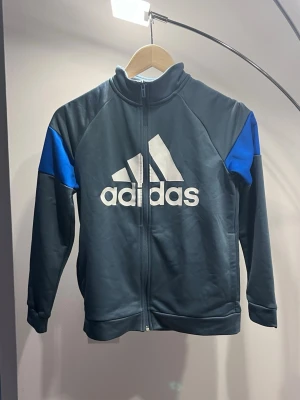 Mörkblå Adidas ziptröja med blå detaljer - Sportig ziptröja från Adidas i mörkblått med blå detaljer på axlarna och stort vitt Adidas-tryck på bröstet. Tröjan har hög krage, hel dragkedja och är tillverkad i ett mjukt, stretchigt material. Perfekt för träning eller chill.