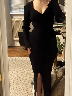 Svart stickad långklänning - Svart stickad långklänning med v-ringad urringning och lång ärm. Klänningen har en figurnära passform och en slits framtill som ger en snygg detalj. Perfekt för dig som gillar en elegant och stilren look.