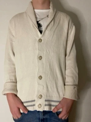 Krämvit kofta från Tommy Hilfiger - Snygg krämvit kofta från Tommy Hilfiger med stora knappar framtill och bred krage. Koftan har ribbstickade muddar och diskreta ränder i nederkanten. Perfekt för lager-på-lager och chill stil.