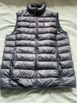 Svart dunväst från Adidas - Snygg svart dunväst från Adidas med klassisk logga på bröstet. Västen har dragkedja framtill, två sidofickor med dragkedja och hög krage. Quiltad design med horisontella sömmar och sportig look. Perfekt för lager-på-lager och streetwear.