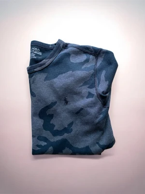 Mörk camo sweatshirt från Polo Ralph Lauren - Snygg sweatshirt i mörkgrått och svart camomönster från Polo Ralph Lauren. Tröjan har rund halsringning, långa ärmar och diskret broderad logga på bröstet. Perfekt för en avslappnad streetwear-look.