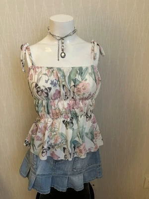 Blommig linnetopp med fjärilar - Söt linnetopp med blommigt och fjärilsmönster i pastellfärger. Toppen har knytbara axelband och volang nedtill för en romantisk look. Perfekt för varma dagar och sommarens alla äventyr.