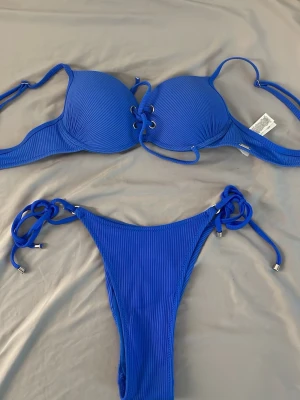 Blå ribbad bikini med snörning - Snygg blå bikini med ribbad struktur och snörning både på toppen och trosorna. Bikinitoppen har vadderade kupor och knyt i ryggen, medan bikinitrosan har knyt i sidorna. Du kan även spänna bhn!jätte fint inför sommaren