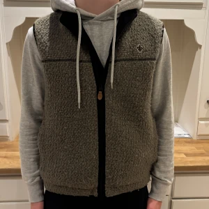 Olivgrön fleeceväst med dragkedja (Morris) - Mysig olivgrön fleeceväst med hög krage och hel dragkedja framtill. Västen har en broderad detalj på bröstet och svarta kantband. Perfekt att slänga över en hoodie för extra värme och stil.