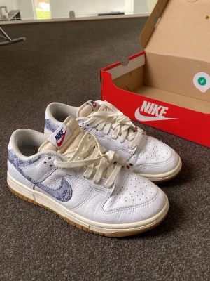 Nike Dunk Low vit/blå  - Nike Dunk Low sneakers i vitt läder med blått mönstrat swoosh och häl. Klassisk siluett med beige yttersula och vita skosnören. Röd och blå Nike-logga på plösen och blå broderad logga på hälen. Perfekt för dig som gillar stilrena men unika detaljer.