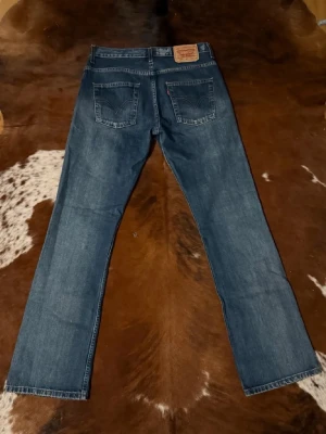 Levi's Bootcut blå jeans - Klassiska blå jeans från Levi's bootcut och fem fickor. Jeansen har en mörk tvätt och tydliga sömmar, samt den ikoniska läderlappen bak i midjan. Tillverkade i slitstark denim, perfekta för en avslappnad och tidlös stil. Storlek W31 L34