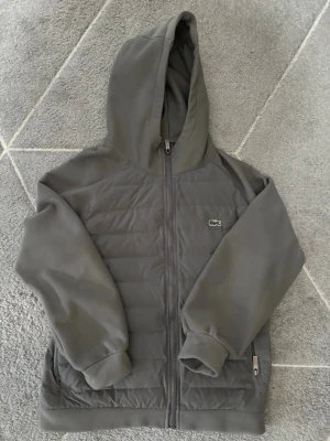 Grå Cardigan från Lacoste med dragkedja - Priset kan diskuteras! Snygg grå Cardigan från Lacoste med dragkedja framtill och klassisk krokodil-logga på bröstet. Hoodien har quiltad framsida, mjuka muddar och fickor med dragkedja. Perfekt för dig som gillar stilren och sportig look.