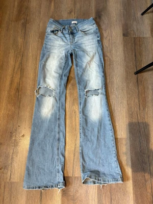 Ljusblå slitna bootcut jeans - Säljer ett par ljusblå bootcut jeans med slitningar och hål över knäna. Jeansen har klassisk femficksdesign, låg midja och råa kanter vid bensluten. Perfekta för en avslappnad och trendig look. Slitna längst nere vid benen bak! Annars inga defekter alls!💓🌸