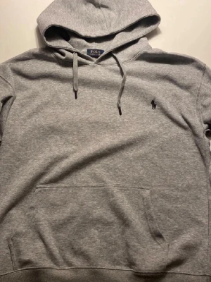 Grå hoodie från Polo Ralph Lauren - Klassisk grå hoodie från Polo Ralph Lauren med broderad logga på bröstet. Hoodien har huva med dragsko, stor magficka och ribbade muddar. Tillverkad i mjuk bomullsblandning som känns skön mot huden. Perfekt för en avslappnad och stilren look.