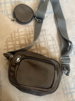 Grå axelväska med extra myntficka - Snygg grå axelväska i slätt material med bred justerbar axelrem och flera dragkedjefack. Väskan har en matchande rund myntficka som sitter fast i remmen. Perfekt för dig som vill ha koll på småsaker och ändå se stilren ut.