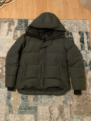 Svart pufferjacka från canada goose - Säljer en svart pufferjacka  med stor huva och diskret grå logotyp på ärmen. Jackan har breda kanaler, dragkedja med vindslå och muddar vid ärmsluten. Perfekt för kalla vinterdagar och riktigt snygg till streetwear-stilen. Men passar till många olika stiler. Jackan är stilren och passar perfekt nu till kalla morgonar och kvällar, den håller dig både varm och bekväm. Den har några ”defekter” vilket jag kan berätta mer om i pm. Påse ingår. Pris kan såklart diskuteras!
