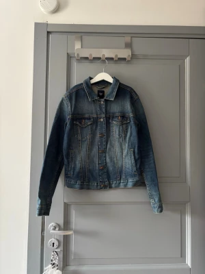 Jeansjacka  - Snygg blå jeansjacka från GAP med klassisk passform. Jackan har två bröstfickor med lock och knappar, samt knappar framtill. Materialet är denim i 100% bomull och jackan har en lätt tvättad look. Perfekt för en cool och avslappnad stil.