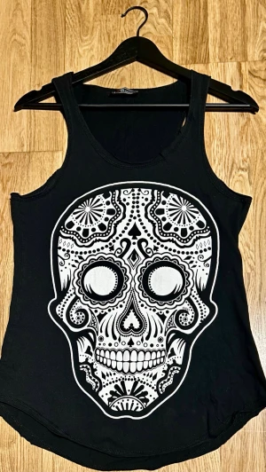 Svart linnetopp med döskallemönster - Cool svart linnetopp från I&S Collection med ett stort, detaljerat vitt döskallemönster på framsidan. Perfekt för dig som gillar edgy och unik stil. Toppens form är klassisk med breda axelband och rund halsringning.                                      Mått: • Byst (armhåla–armhåla): ca 42 cm • Längd: ca 67 cm