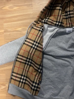 Grå hoodie från Burberry med rutigt foder - Säljer en grå zip-hoodie från Burberry med det klassiska beige, svart och rödrutiga fodret i huvan och insidan. Hoodien har dragkedja, fickor fram och justerbar huva med snören. Perfekt för dig som vill ha en stilren men ändå lyxig look. Priset kan diskuteras vid snabb affär