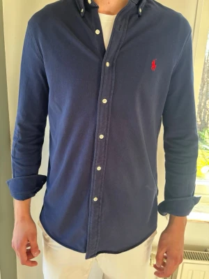 Ralph Lauren Skjorta Featherweight Mesh | Marinblå M  - Sjukt snygg marinblå skjorta från Ralph Lauren. Perfekt mjuk kvalitet! Modellen är 189 cm.  En otroligt fin garderobsfavorit. 👕 Märke & Modell: Ralph Lauren - Featherweight Mesh 📏 Storlek & Passform: M - Slim fit/Custom fit 🧵 Material: 100% bomull 📐 Plaggets mått: Bröst 50 cm, längd 70 cm, ärm 46 cm ✅ Skick: 9/10, inga defekter, mycket mjuk ✨ Detaljer: Röd logga, knappkrage, vita knappar 💰 Nypris: ca 1500 kr ‼️ Skickas inom 24h ‼️