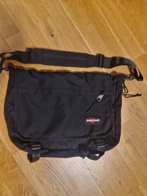 Svart axelväska från Eastpak - Säljer en svart axelväska från Eastpak med justerbar axelrem och vadderad axelkudde. Väskan har ett stort huvudfack, en ficka med dragkedja på framsidan och spännen för extra säkerhet. Perfekt för skola eller cityhäng.