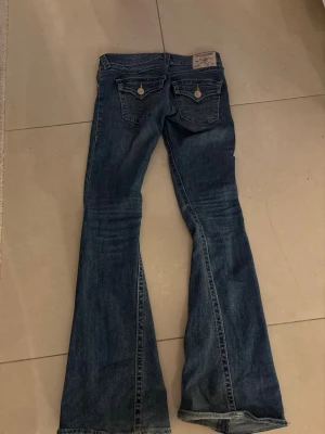 Blå low waist bootcut jeans från true religion - Säljer ett par blå bootcut jeans från true religion med klassiska bakfickor med lock och knappar. Jeansen har låg midja.