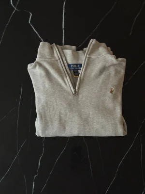 Grå halvzip från Polo Ralph Lauren - Klassisk ljusgrå tröja från Polo Ralph Lauren med halv dragkedja och broderad logga på bröstet. Tröjan har ribbade muddar vid ärmslut och nederkant samt hög krage. Perfekt för en avslappnad men stilren look. Längd 62cm bredd 45cm
