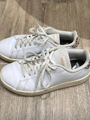 Vita Adidas sneakers med leoparddetalj - Snygga vita Adidas sneakers i läder med klassisk låg profil och leopardmönstrad detalj på hälen. Skorna har vita snören, diskreta perforeringar på sidan och Adidas-logga i guld på plösen. Perfekta för dig som vill ha en unik twist på dina sneakers.