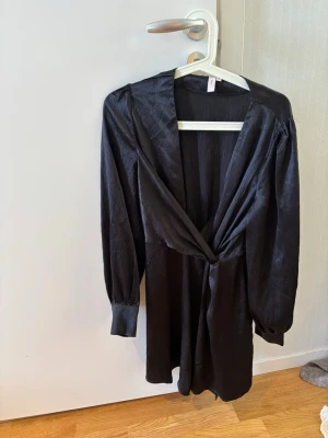 Svart omlottklänning med v-ringning - Stilren svart omlottklänning med djup v-ringning och långa ärmar. Klänningen har en lätt glansig yta och markerad midja som ger en snygg siluett. Perfekt för dig som gillar en enkel men elegant look. Storlek 36 