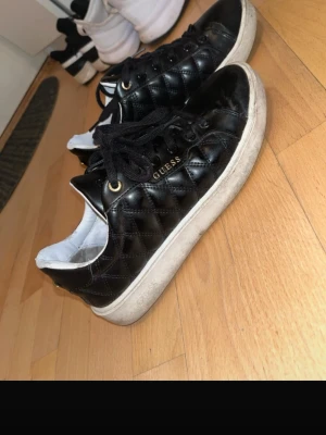 Svarta quiltade sneakers från Guess - Snygga svarta sneakers från Guess med quiltad design och guldfärgade detaljer. Skorna har vita sulor, svarta skosnören och Guess-logga på sidan och plösen. Tillverkade i konstläder med vit insida för en fräsch kontrast. 