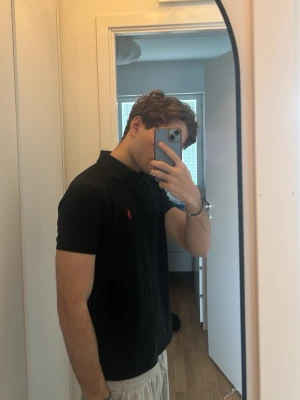 Svart piké från Polo Ralph Lauren - Klassisk svart pikétröja från Polo Ralph Lauren med röd broderad logga på bröstet. Tröjan har korta ärmar, krage och knappar vid halsen. Tillverkad i mjuk bomull som är skön att bära och passar till det mesta.