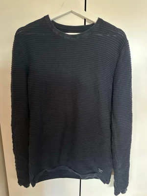 Mörkblå ribbad tröja | Jack & Jones - Mörkblå långärmad tröja från Jack & Jones med ribbad struktur och rund hals. Tröjan har en enkel och stilren design, perfekt för en avslappnad look. Materialet är mjukt och bekvämt, och den har en liten diskret etikett nertill. Dock finns det tre små hål på baksidan samt ett litet hål på ärmen därav priset. Storlek M men är stor i sin storlek, så skulle säga kring L.