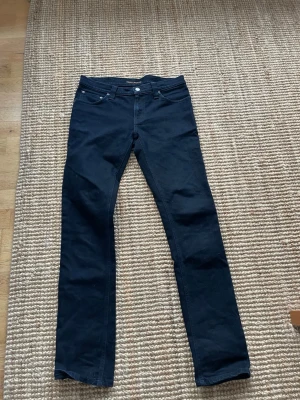 Svarta jeans från Nudie Jeans - Säljer dessa sjukt Snygga svarta jeans i från Nudie Jeans med raka ben. Jeansen har silverfärgade nitar och knapp, samt bälteshällor. Materialet är mjukt och stretchigt, perfekt för en avslappnad men stilren look.