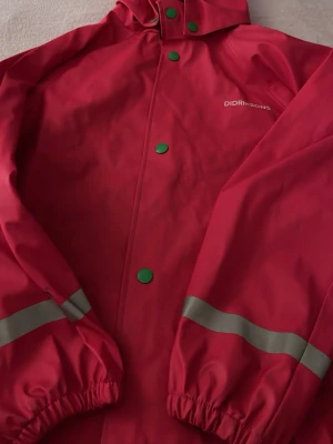 Röd regnjacka från Didriksons - Snygg röd regnjacka från Didriksons med gröna tryckknappar och reflexdetaljer på ärmarna. Jackan har huva och elastiska muddar vid ärmsluten. Perfekt för regniga dagar och enkel att matcha med andra plagg.