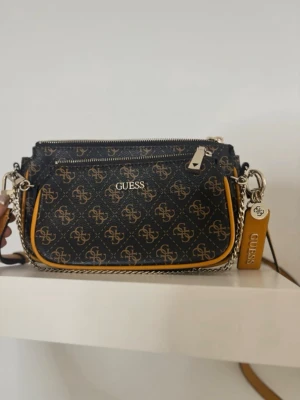 Svart och brun handväska från Guess - Snygg handväska från Guess i svart och brunt med logomönster över hela väskan. Väskan har guldfärgade detaljer, kedjerem och en dekorativ tag med Guess-logga. Tillverkad i konstläder och har dragkedja upptill.