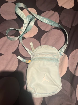 Ljusblå axelväska från Jordan - Snygg ljusblå axelväska från Jordan med justerbar axelrem och dragkedja framtill. Väskan har en minimalistisk design med det klassiska Jumpman-loggan broderad på framsidan. Perfekt för att bära det viktigaste och enkel att matcha med olika outfits.