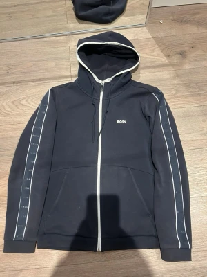 Svart hoodie med dragkedja från BOSS - Snygg svart hoodie från BOSS med vit dragkedja och vita detaljer längs ärmarna. Huva med dragsko och fickor framtill. BOSS-logga på bröstet och diskreta loggor längs ärmarna. Perfekt för en chill och stilren look.