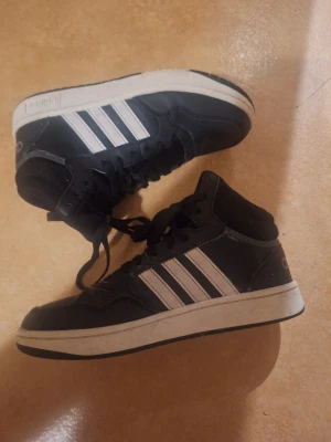 Svarta Adidas high top sneakers - Snygga svarta high top sneakers från Adidas med klassiska vita ränder på sidorna och vit sula. Skorna har snörning och en sportig look som passar perfekt till streetwear. Tillverkade i syntetmaterial och textil för en bekväm känsla.