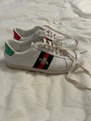 Gucci Ace vita sneakers  - Säljer ett par vita Gucci Ace sneakers i läder med klassiska gröna och röda ränder på sidan och en broderad guldfärgad bi. Skorna har grönt och rött parti på hälen och vita snören. Perfekta för dig som vill ha en ikonisk och trendig look.