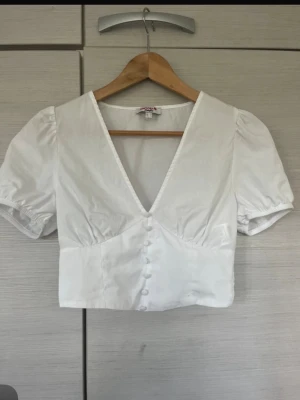 Vit kort blus med puffärm - Trendig vit blus med djup v-ringning och puffiga korta ärmar. Knäppning framtill med tygklädda knappar och smock baktill i midjan för en snygg passform. 