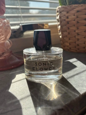 Sonic Flower parfym från Room 1015 - Sonic Flower Eau de Parfum från Room 1015 i en transparent glasflaska med svart, multifacetterat lock. Flaskan har en modern och stilren design med tydlig text på framsidan. Perfekt för dig som gillar unika och fräscha dofter. 50ml, mer än hälften kvar