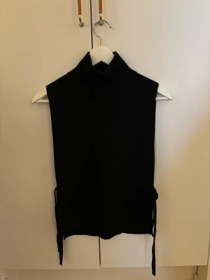 Svart stickad väst från ARKET - Svart stickad väst från ARKET med polokrage och knytband i sidorna. Västen är i mjuk merinoull och har en clean, minimalistisk look som passar perfekt över en skjorta eller tröja.