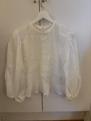 Vit blus med puffärm och struktur - Stilren vit blus med rund halsringning och puffiga långärmar. Framsidan har ett snyggt strukturerat mönster och tyget är lätt och något transparent. Blusen har knappstängning bak och resår vid ärmsluten för en trendig look.