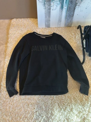 Svart sweatshirt från Calvin Klein - Snygg svart sweatshirt från Calvin Klein Jeans med stor logga framtill. Tröjan har rund halsringning och långa ärmar. Tillverkad i mjukt material som passar perfekt till en avslappnad stil.