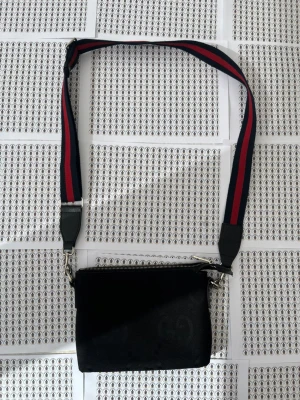 Svart axelväska från Gci - Snygg svart axelväska från Gci med diskret G-mönster i tyget. Väskan har en bred axelrem i tyg med röd och marinblå rand, samt silverfärgade metalldetaljer och dragkedja upptill. Perfekt för dig som gillar stilrena accessoarer med lyxig känsla.