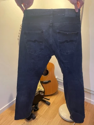 Nudie Jeans Grim tim  - Säljer ett par mörkblå jeans från Nudie Jeans med klassisk femficksmodell och raka ben. Jeansen har Nudie Jeans karakteristiska broderi på bakfickorna och är tillverkade i denim. Perfekta för en avslappnad och stilren look.