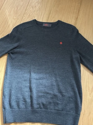 Mörkgrå stickad tröja från Sunwill - Mörkgrå stickad tröja från Morris med rund hals och diskret röd logotyp broderad på bröstet. Tröjan har långa ärmar och ribbade muddar vid ärmslut och nederkant. Materialet är mjukt och tunt, perfekt för lager-på-lager och snyggt till jeans.
