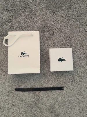 Svart armband från Lacoste - Snyggt svart armband från Lacoste med modern, stilren design. Armbandet har en länkad struktur i metall och kommer med originalförpackning och påse. Perfekt accessoar för dig som gillar minimalistisk och sportig stil.