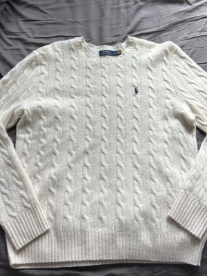Vit kabelstickad tröja Polo Ralph Lauren - Vit kabelstickad tröja från Polo Ralph Lauren med rund halsringning och broderad logga på bröstet. Tröjan har långa ärmar och ribbstickade muddar samt nederkant. Perfekt för en stilren och klassisk look.