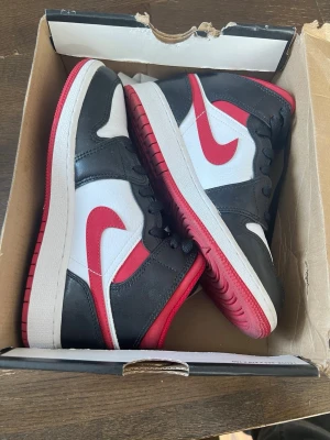 Nike Air Jordan 1 Mid Svart/Röd/Vit - Snygga Nike Air Jordan 1 Mid sneakers i svart, vitt och rött. Skorna har klassisk röd sula, vit ovandel med svarta och röda detaljer samt den ikoniska swoosh-loggan i rött. Tillverkade i läder och textil för en clean och sportig look. 