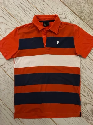 Randig Peak Performance piké - Snygg randig pikétröja från Peak Performance i rött, marinblått och vitt. Skjortan har klassisk krage, knappar framtill och breda horisontella ränder. Perfekt för en sportig och avslappnad stil.