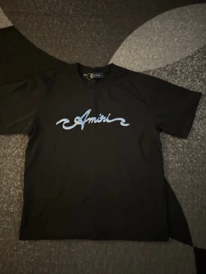 Amiri t-shirt - Säljer en svart t-shirt från Amiri med ett stort blått logotryck över bröstet. Klassisk rak passform och rund halsringning. Perfekt för dig som gillar streetwear och vill ha något stilrent men ändå med attityd. Storlek M. Aldrig använd.