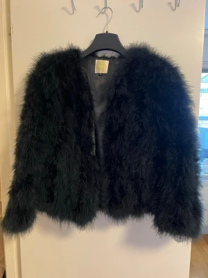 Svart fluffig pälsjacka från Pello bello - Snygg svart fjäderjacka från Pello bello med fluffig och voluminös look. Jackan har rundad halsringning, helsvart foder och stängs framtill. Perfekt för dig som vill ha en statementjacka med lyxig känsla och trendig siluett.