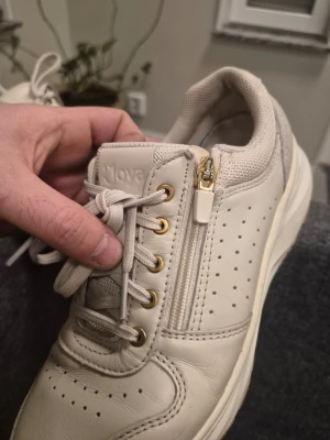 Beige sneakers från Joya med gulddetaljer 37 ⅔ - Snygga beige sneakers från Joya med guldfärgade detaljer och dragkedja på sidan. Skorna har perforerat läder, snörning och vadderad häl för extra komfort. Perfekta för dig som vill ha både stil och funktion i vardagen.