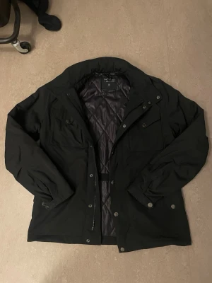 Svart peak performance fieldjacket - Stilren svart fieldjacket från peak performance med två bröstfickor med lock och knappar, samt dragkedja och tryckknappar framtill. Jackan har en hög krage och diskret logotyp på ärmen. Perfekt för blåsiga dagar och enkel att matcha.